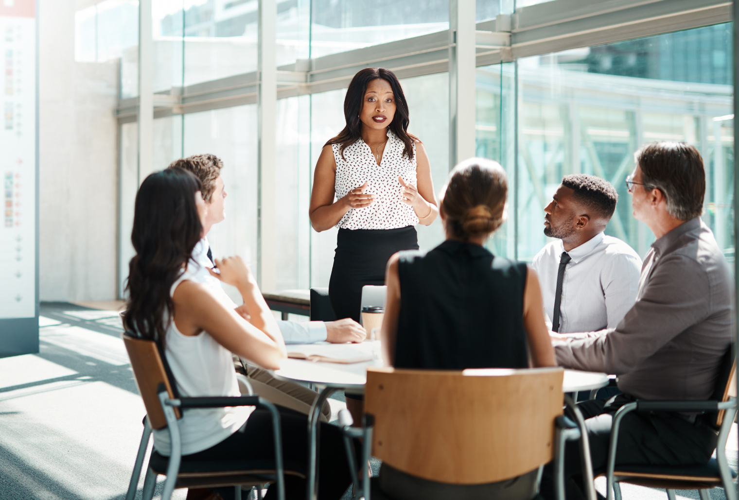 A group of people, men and women, having a meeting on a nice day by big glass walls.