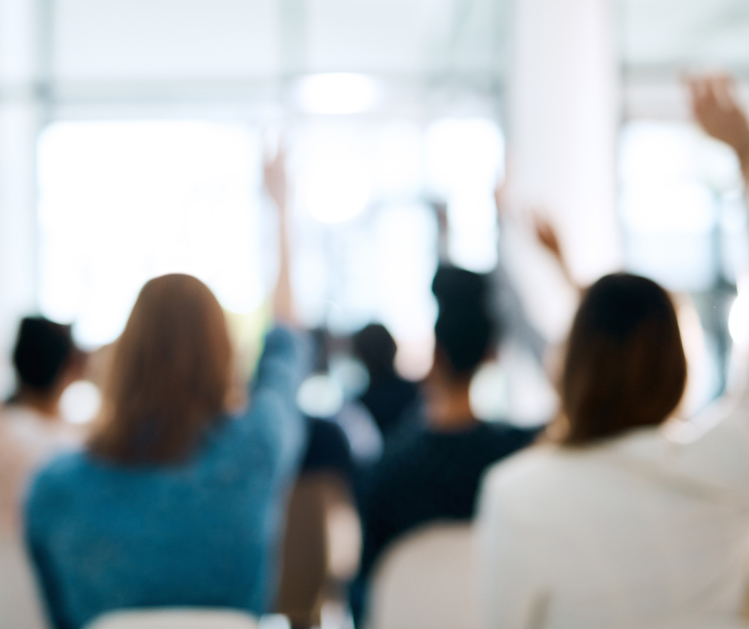 Several people raising their hands in a blurred picture of a business meeting.